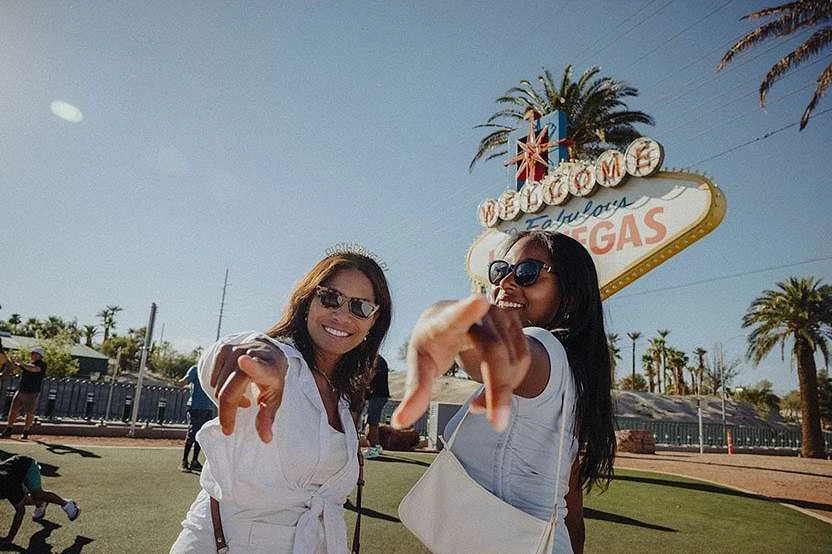 Two smiling women pose in front of the 'Welcome to Fabulous Las Vegas' sign, both wearing sunglasses and casual summer outfits on a sunny day.