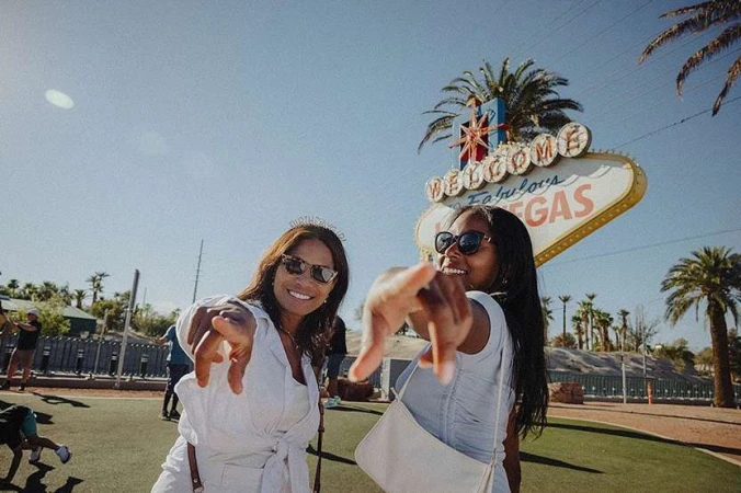 Two smiling women pose in front of the 'Welcome to Fabulous Las Vegas' sign, both wearing sunglasses and casual summer outfits on a sunny day.