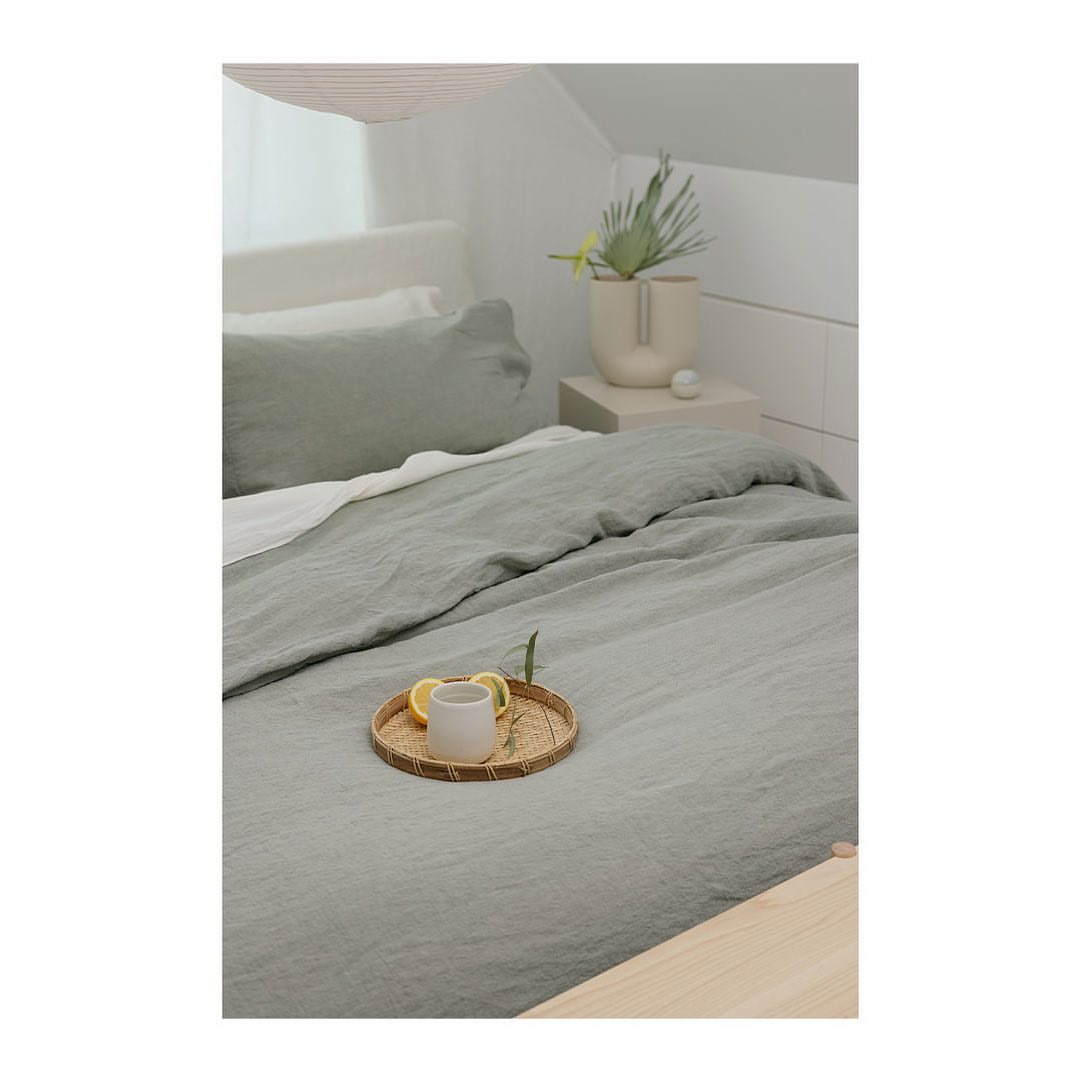 Neutral bedroom interior featuring a green linen duvet, decorative tray with mug and lemons, and a bedside table with plants.