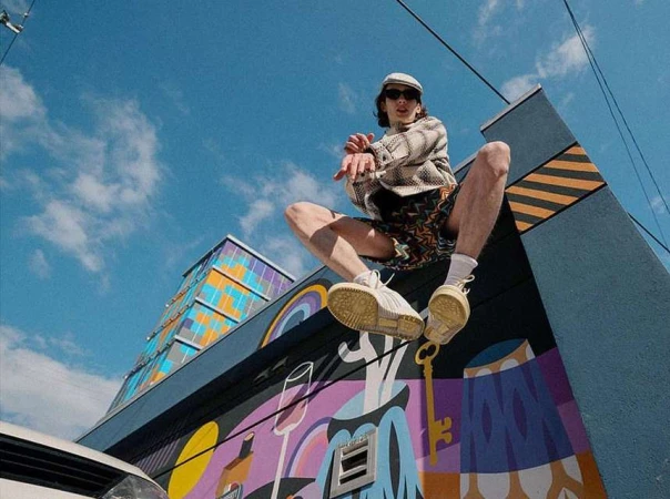 Man with sunglasses and hat sitting on a building with a mural, wearing patterned shorts and sneakers against a sky background.