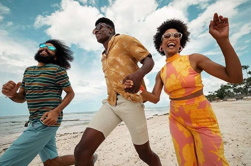 Three adults happily run on a sunny beach. The woman in the center wears an orange floral outfit and sunglasses, and the others are smiling.
