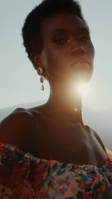 Portrait of a stylish Black woman in a floral dress against a backdrop of mountains, illuminated by the sun, accessorized with earrings.