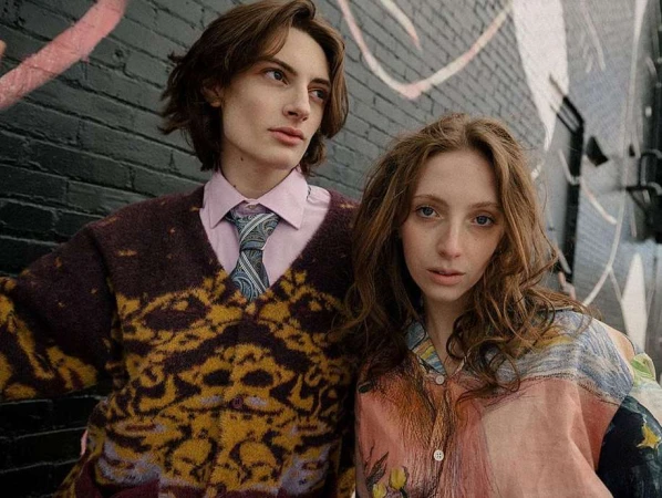 Two models posing outdoors against a dark brick wall with colorful painted abstract graffiti markings.