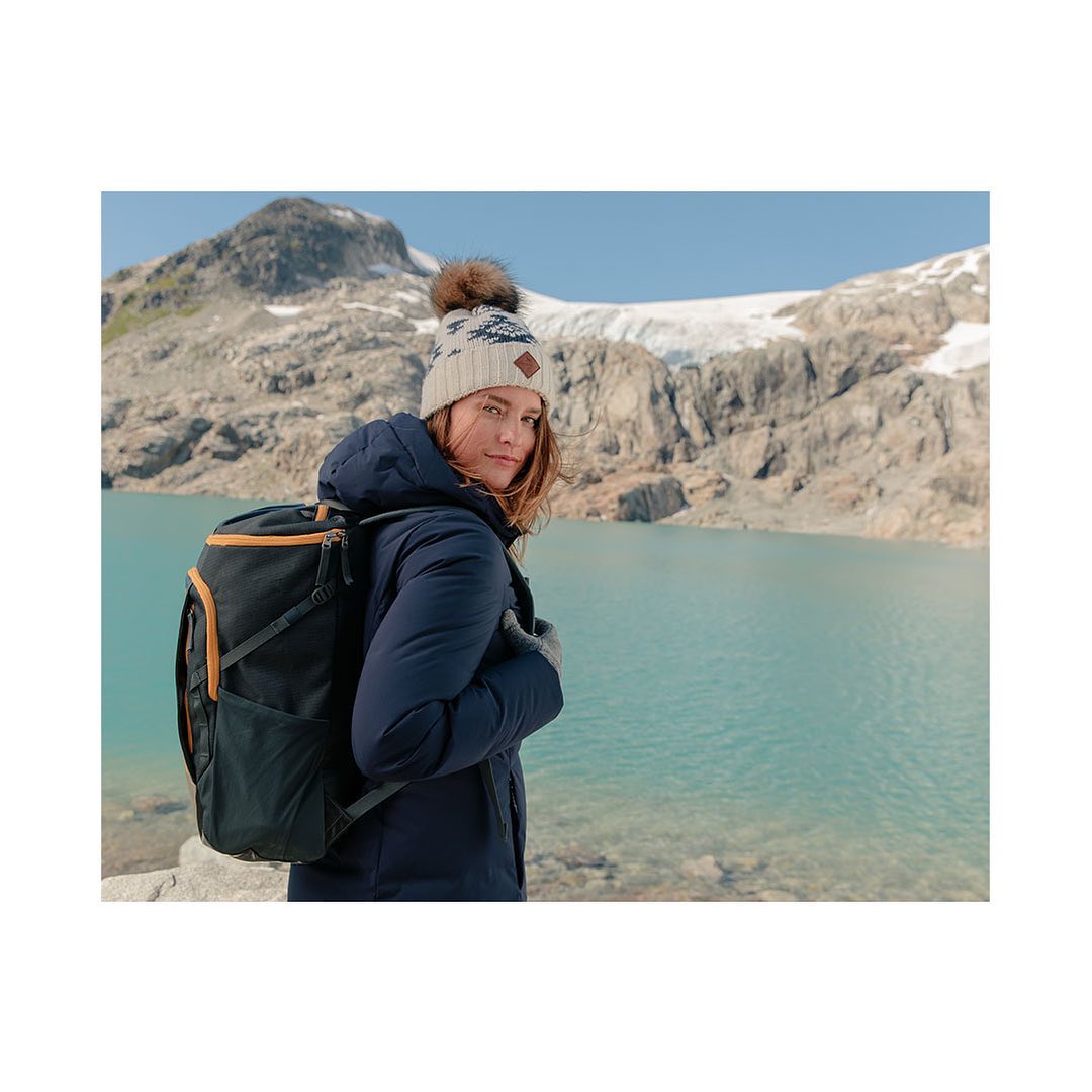A woman with a backpack, wearing a hat and winter coat, stands against a background of mountains and a turquoise glacial lake on a sunny day.