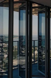 Modern glass folding doors open to a high-rise balcony overlooking a coastal city skyline and ocean.