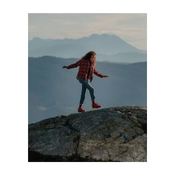 A woman balancing on a rocky outcrop wearing a plaid shirt and blue jeans against a mountain backdrop.