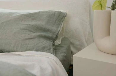 Close-up of a made bed with sage green linen pillows and white sheets beside a modern cream nightstand and vase