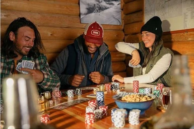 Three friends laughing while playing cards and sitting around a wooden table indoors.