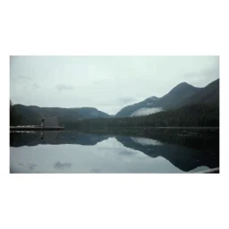 Calm lake reflecting forested mountain peaks and overcast sky under soft light