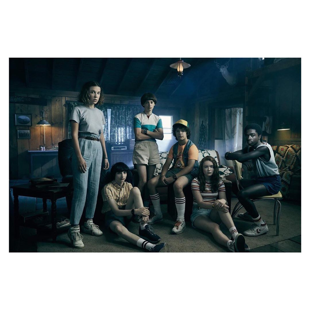 Group shot of six young actors in retro late 20th-century clothing in dimly lit home setting, posing for a promotional image.