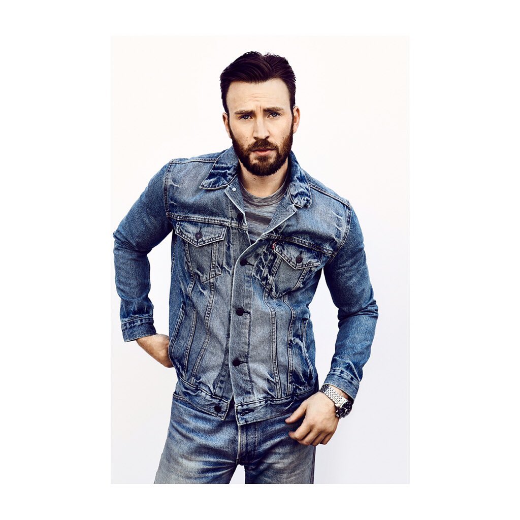 Portrait of a bearded adult male wearing a denim jacket and jeans against a neutral background, hands in his pockets, looking at the camera.