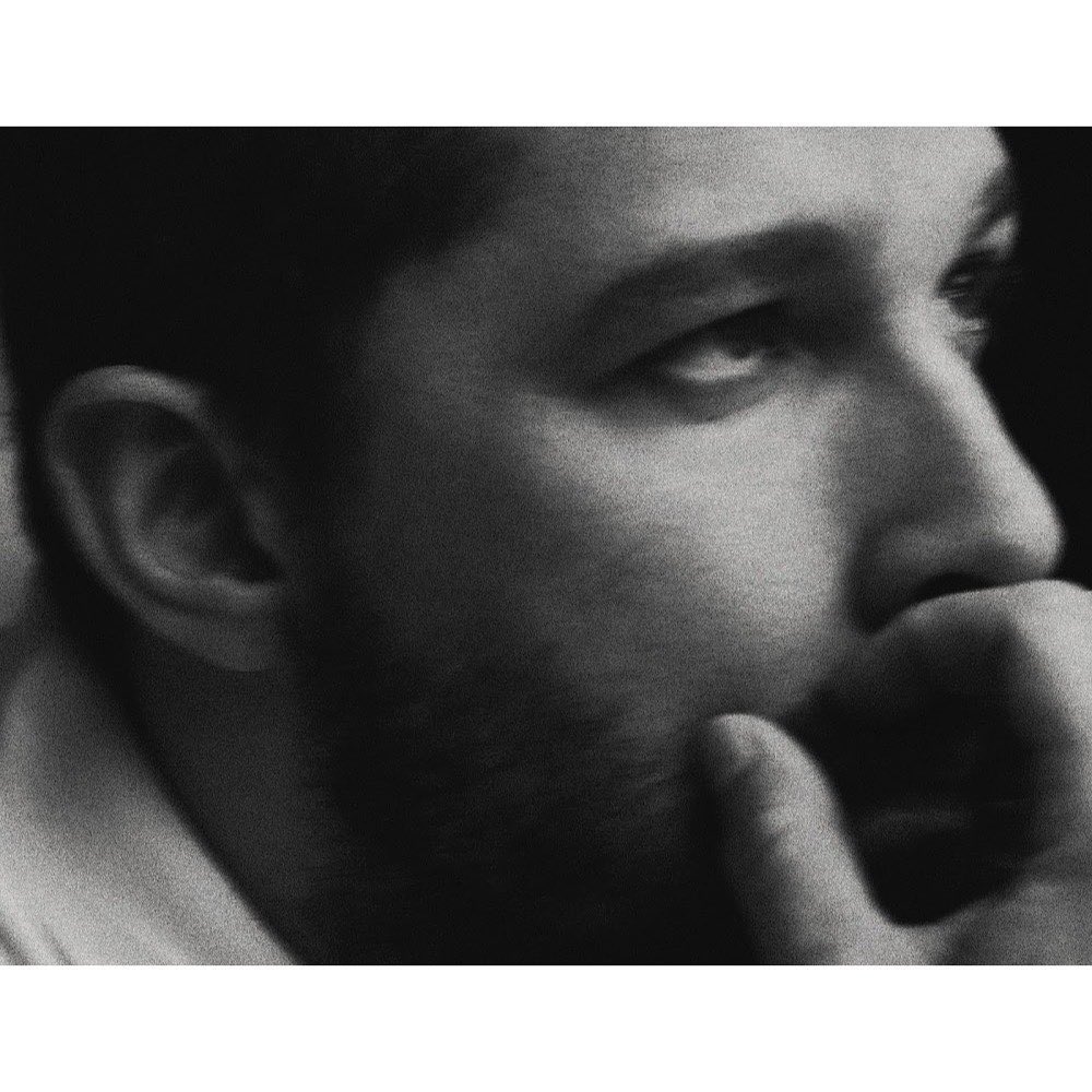 Close-up portrait of an adult male with his hand near his mouth, depicted in black and white with soft, diffused lighting and shallow focus.