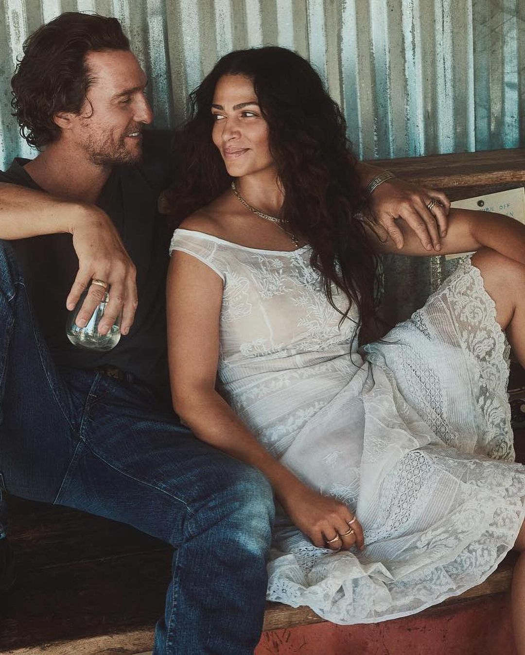 A couple poses together. A smiling man in jeans holds a glass next to a woman in a lacy white dress and long hair, sitting inside.