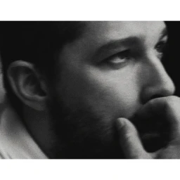 Close-up black and white portrait of a man with dark hair and a beard, resting his hand near his chin.