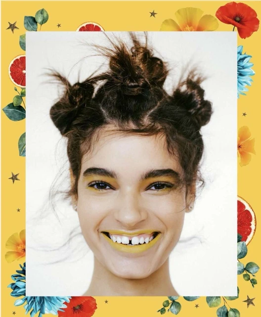 Close-up portrait of a smiling young woman with unique hair buns and yellow makeup framed by a decorative floral and fruit border.