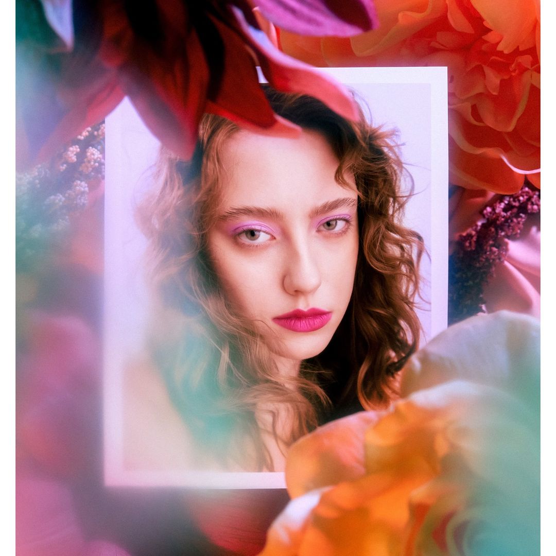 Close-up portrait of a young woman with curly brown hair, pink eyeshadow, and bright lipstick, framed by blurred colorful flower petals.