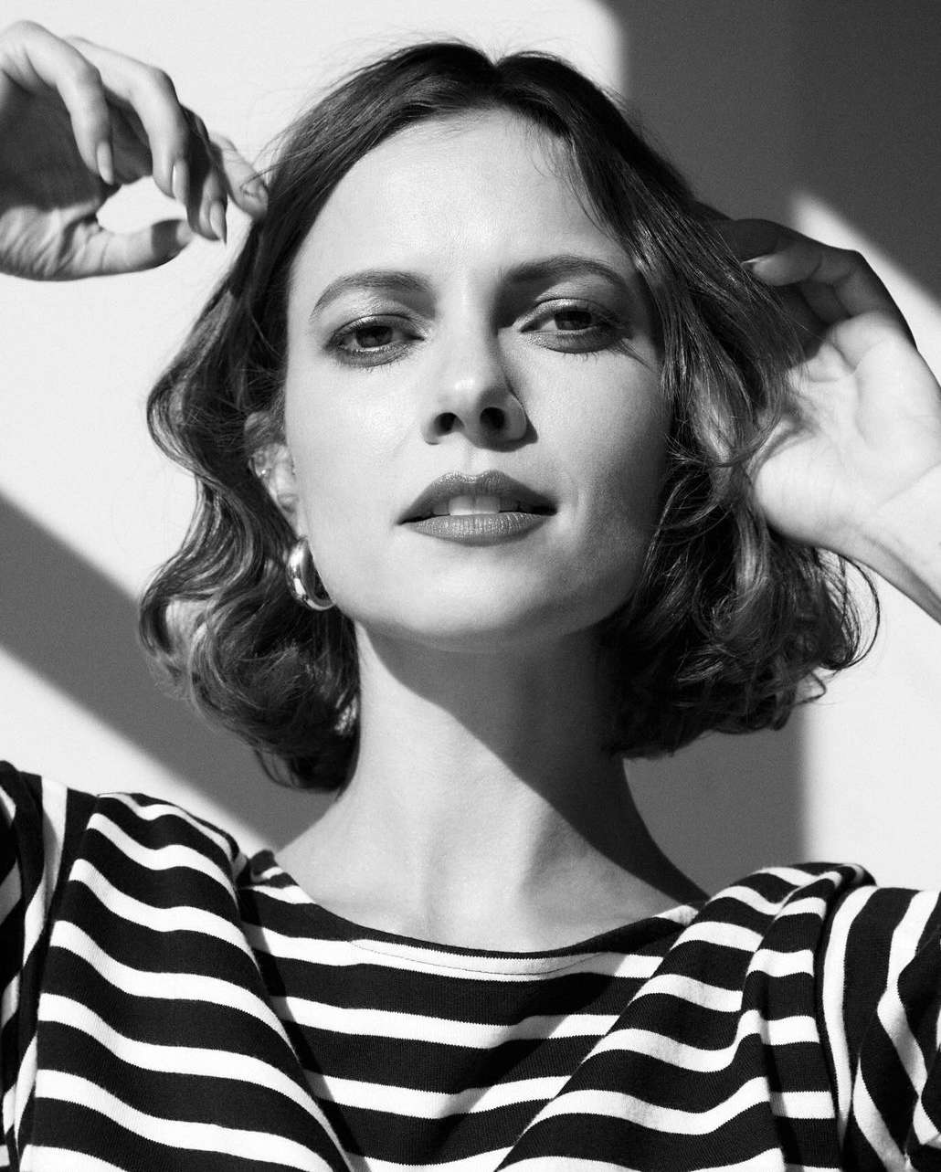 Black and white portrait of a woman with hands near her hair, wearing a striped shirt against a neutral backdrop with soft lighting.