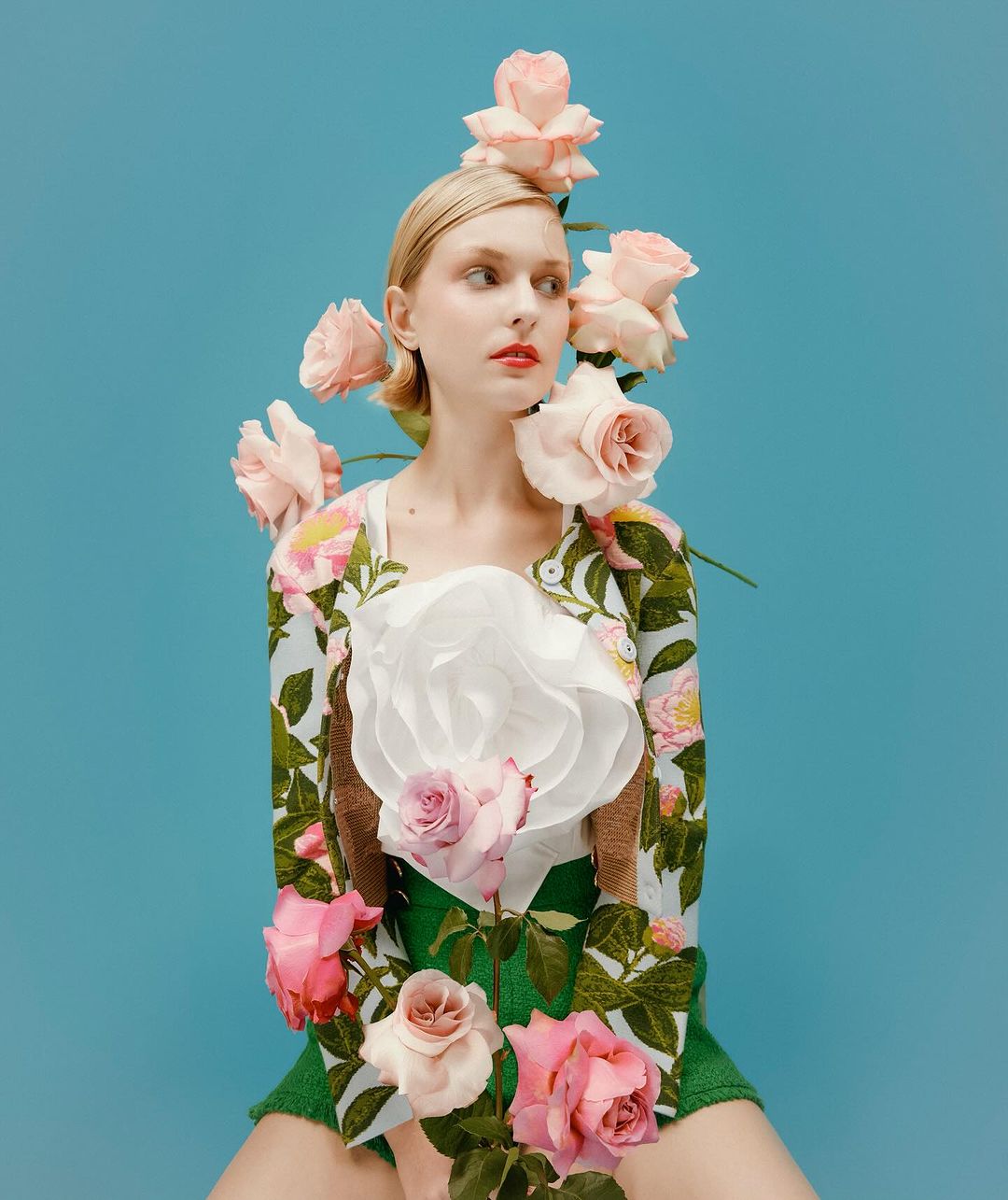 Woman with a pale complexion wears a floral-print top with pink roses and green shorts against a light blue backdrop in a studio setting.