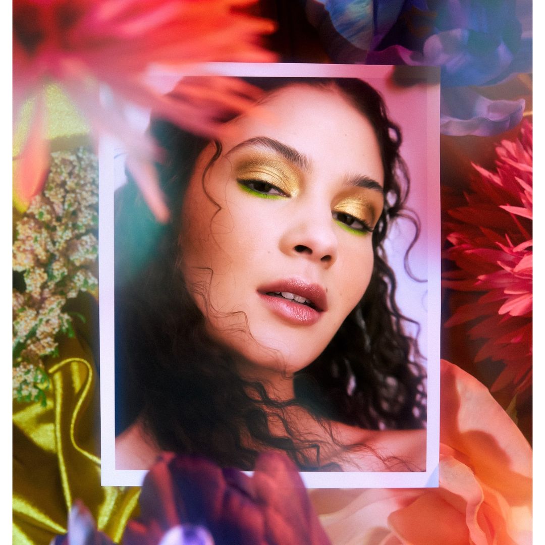 Close-up of a woman with golden eyeshadow and dark, curly hair surrounded by flowers, creating a visually striking beauty portrait.