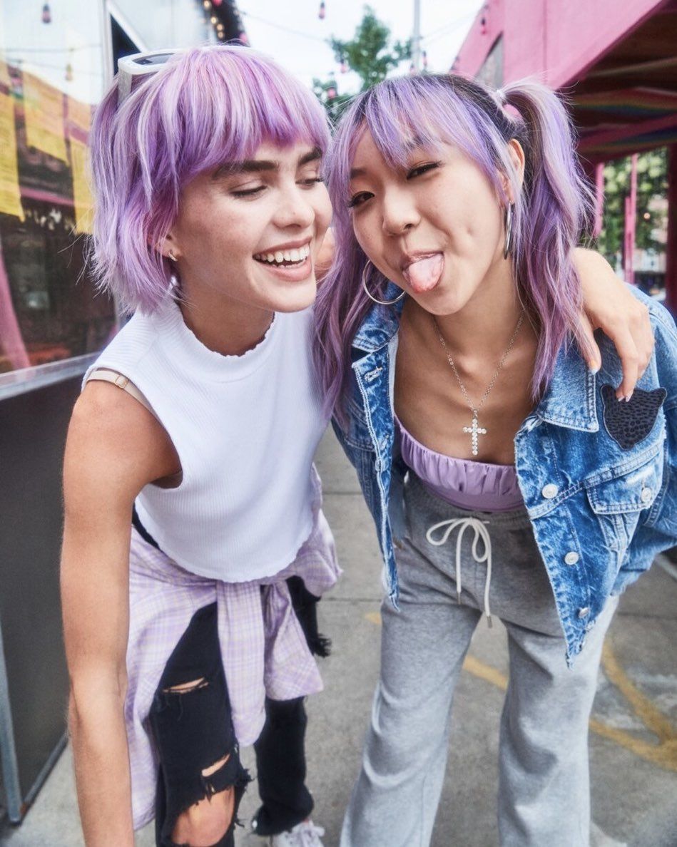 Two young adult women with purple hair pose closely together outdoors, smiling. One woman sticks out her tongue playfully.