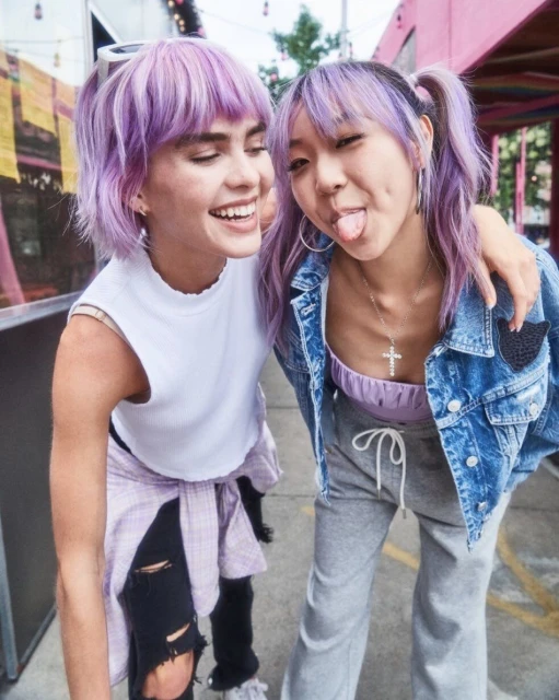 Two young adult women with purple hair pose closely together outdoors, smiling. One woman sticks out her tongue playfully.