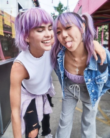 Two young adult women with purple hair pose closely together outdoors, smiling. One woman sticks out her tongue playfully.