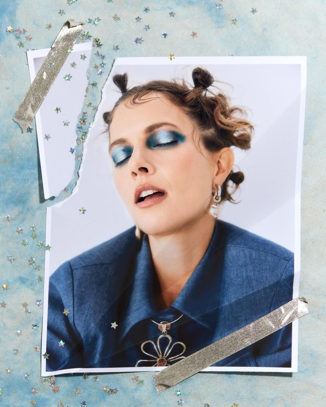 Woman in a photograph with blue eyeshadow, wearing a denim shirt and flower necklace, taped to a glittery blue background.