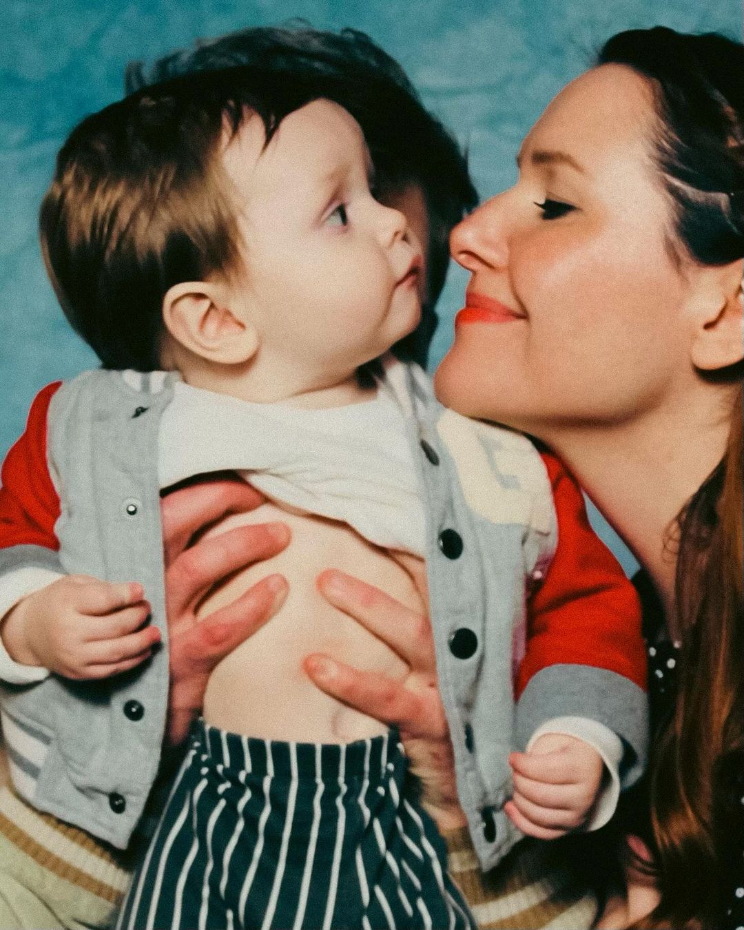 A mother in a polka dot dress smiling at her baby held up by a third person in a red and grey jacket and striped pants in a photo studio.