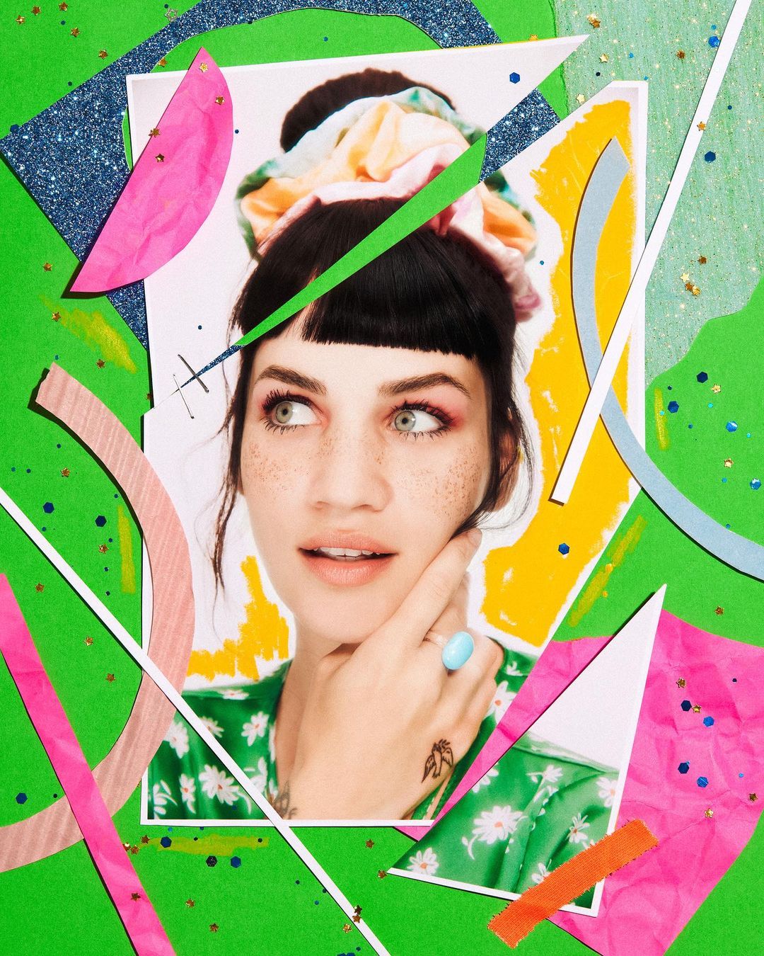 Woman with a hair bun looks off-camera, featuring colorful paper cutouts, a green background, and an eye-catching ring. Beauty shot.