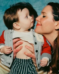 Woman holding a toddler wearing a red and grey varsity jacket and striped trousers against a textured blue backdrop