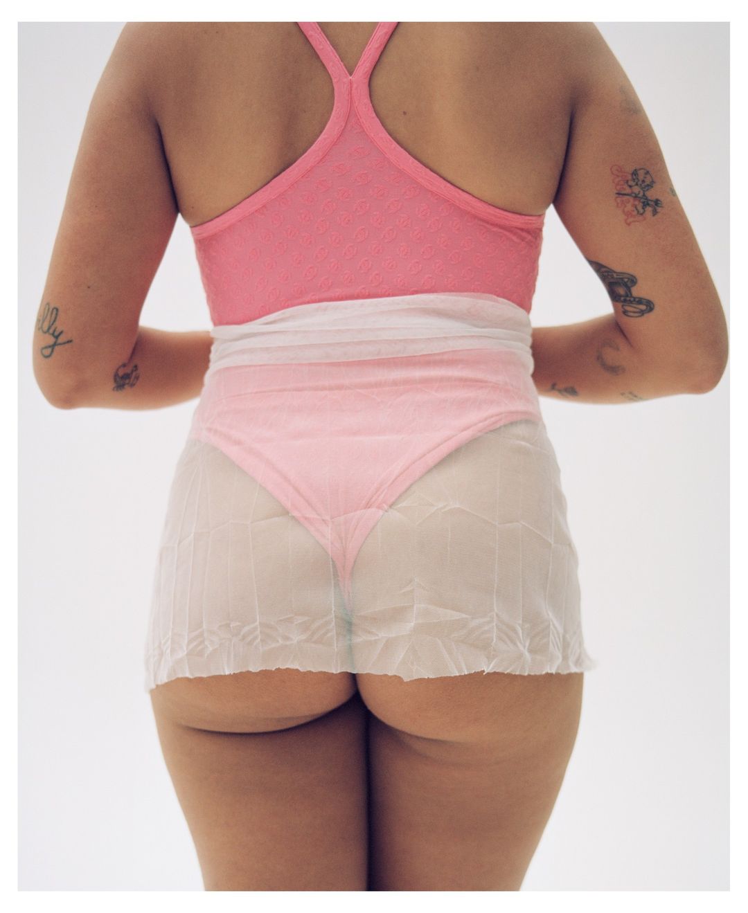 Rear view of a female model wearing a pink top, white skirt, and pink underwear in a photo studio. Tattoos visible on arms.