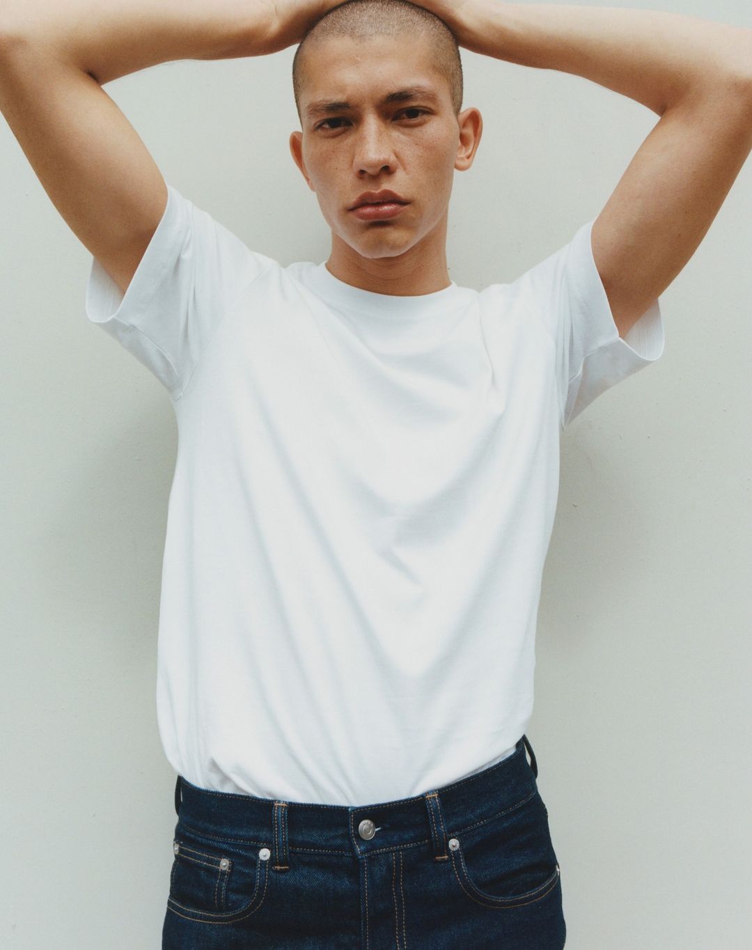 Portrait of a young adult man with a shaved head wearing a white t-shirt and blue jeans, posing with his hands behind his head against a white wall.