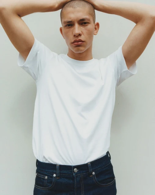 Portrait of a young adult man with a shaved head wearing a white t-shirt and blue jeans, posing with his hands behind his head against a white wall.