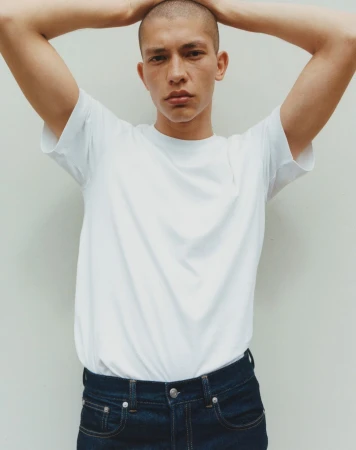 Portrait of a young adult man with a shaved head wearing a white t-shirt and blue jeans, posing with his hands behind his head against a white wall.
