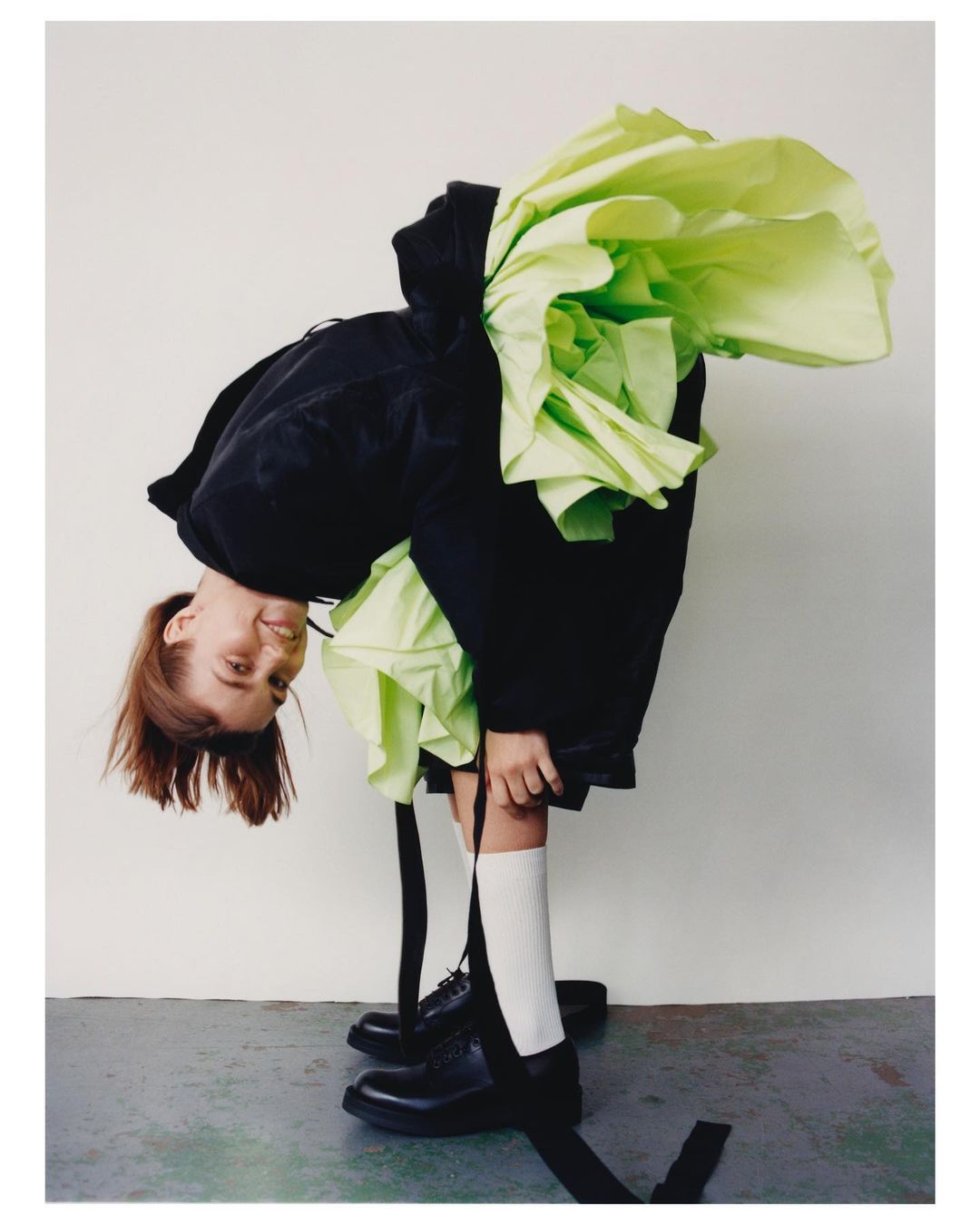 Young person in a black hoodie and skirt with light green tulle bends over, facing downward with a smile, wearing black shoes and white socks.