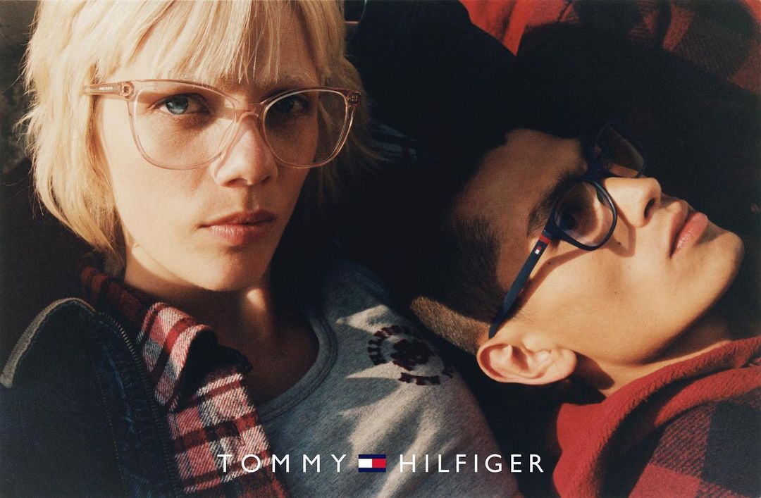 Close-up of a woman with blonde hair and glasses next to a man, both wearing clothing with the Tommy Hilfiger logo.