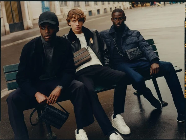Three men sitting together on a green park bench wearing dark jackets and crossbody bags, photographed by Misha Taylor