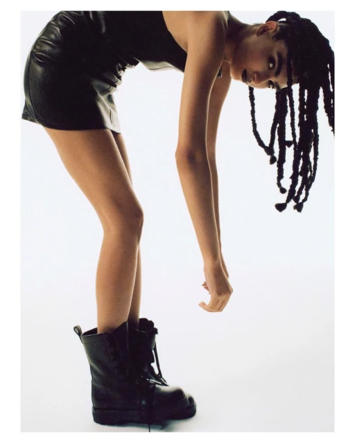Fashionable woman with braids bends over in black mini dress and chunky black boots against a white backdrop in a studio shot.