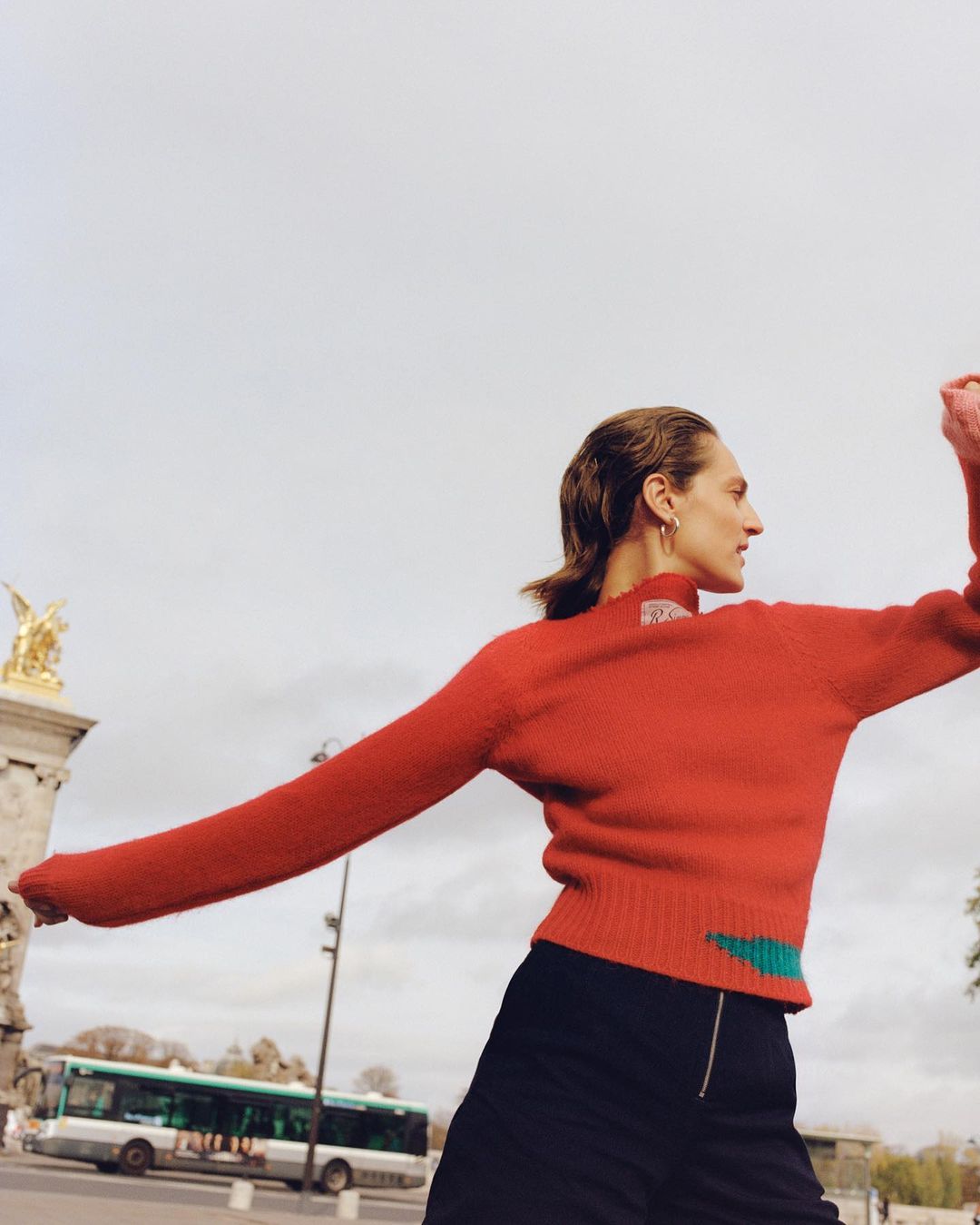 Woman in red sweater and black skirt stands on city street with bus and architecture in background. Fashion shot, overcast day.