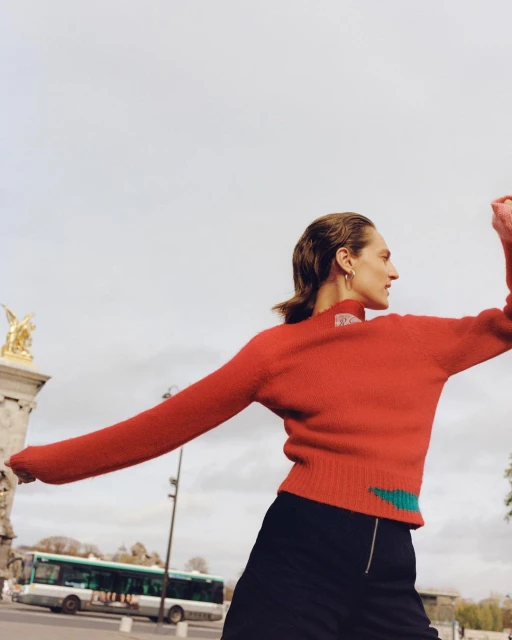 Woman in red sweater and black skirt stands on city street with bus and architecture in background. Fashion shot, overcast day.