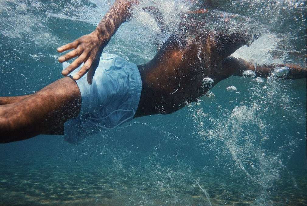 Underwater shot of a man swimming in light blue swim trunks. Bubbles and ripples in the water. Ocean floor visible in the distance.