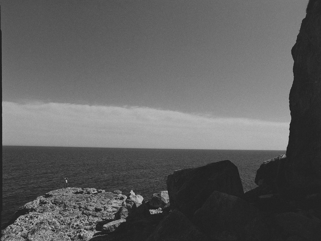 Black and white landscape featuring the ocean, a rocky cliffside, and a distant person on the edge, under an expansive sky.