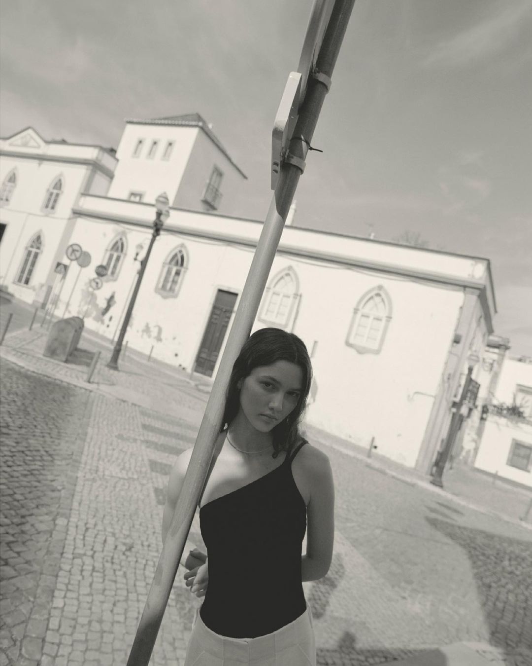 Monochrome portrait of a young woman wearing a one-shoulder top stands on a cobblestone street in front of historic buildings.