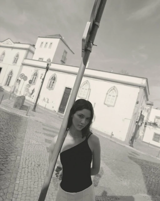 Monochrome portrait of a young woman wearing a one-shoulder top stands on a cobblestone street in front of historic buildings.