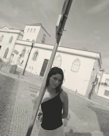 Monochrome portrait of a young woman wearing a one-shoulder top stands on a cobblestone street in front of historic buildings.