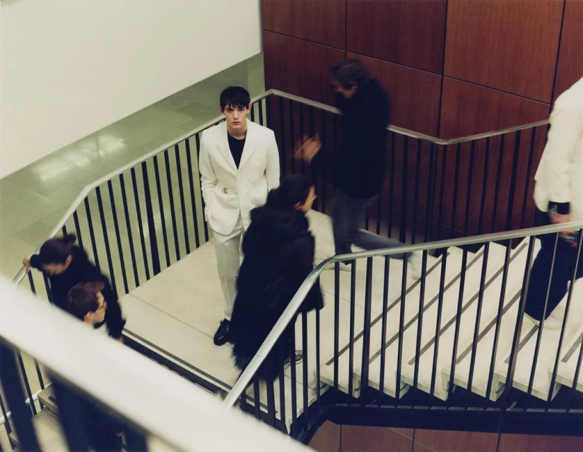 A young man in a white suit stands on a staircase as people ascend and descend. An overhead angle captures the modern, urban scene.