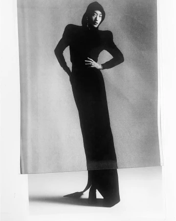 Monochrome fashion portrait of a model wearing a dark hooded outfit with structured shoulders against a seamless studio background.