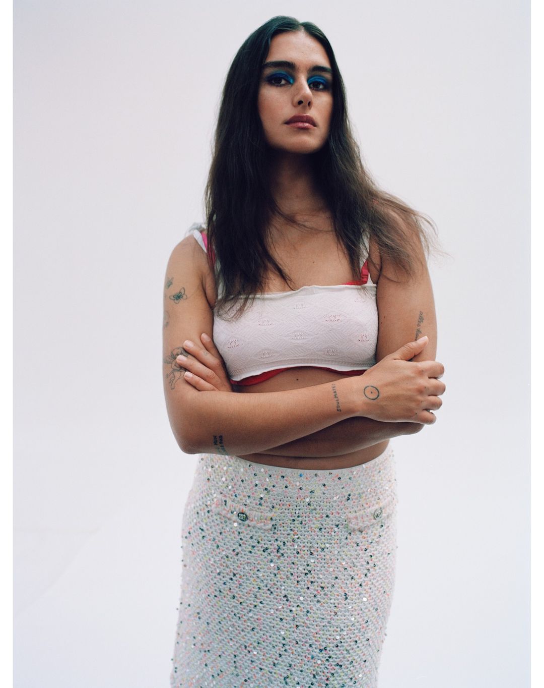 Portrait of a young woman with dark hair and blue eye shadow wearing a white top and skirt against a plain background.