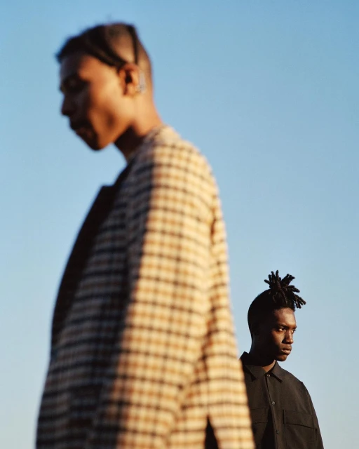 Two African American men in an outdoor setting. One man is out of focus in the foreground, wearing a patterned tan and brown coat. The other is more in focus.