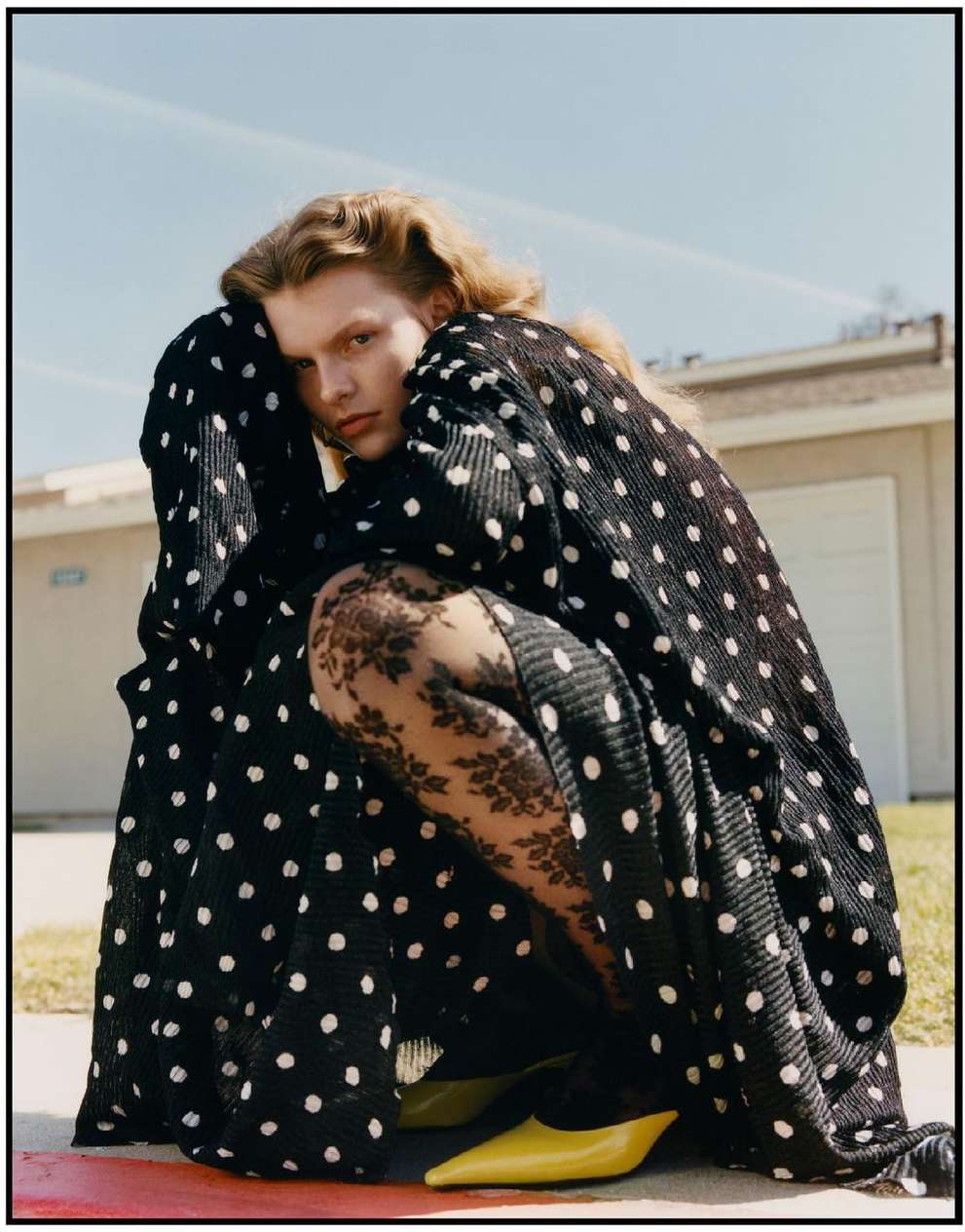 Editorial fashion shot of a young adult woman in a polka dot dress, crouched outside in front of a building on a sunny day.